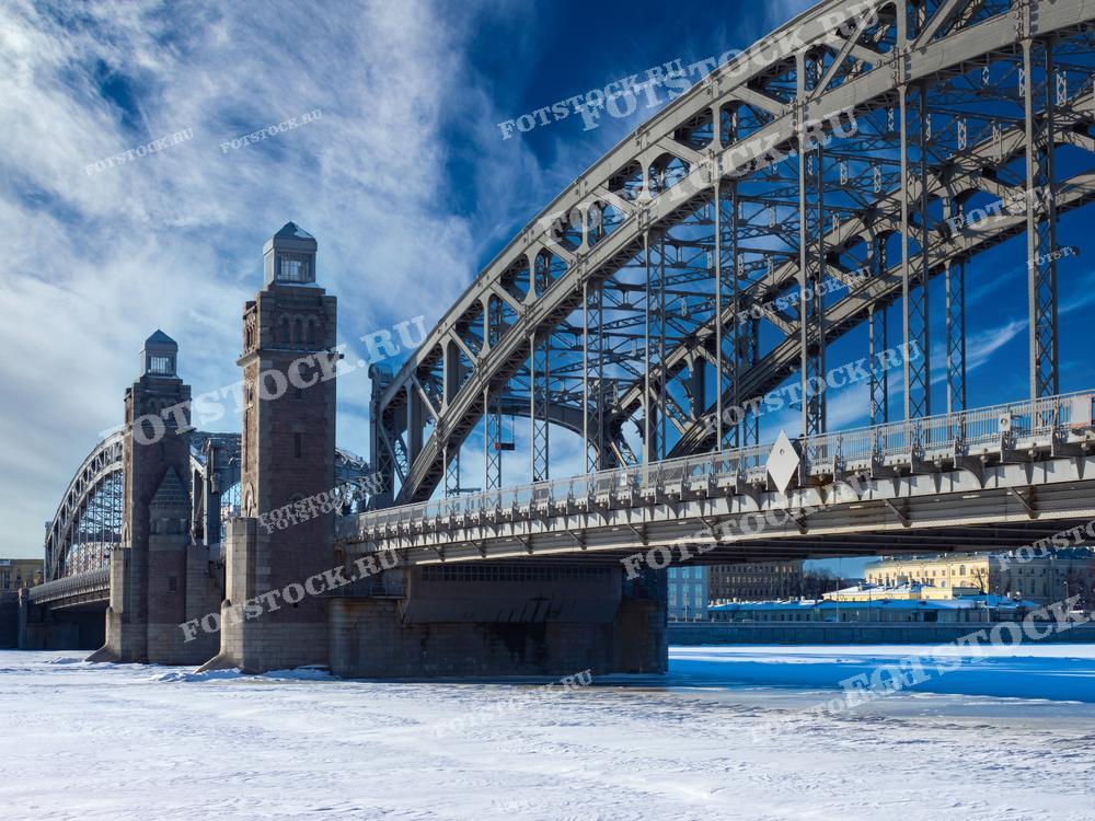 На фотографии изображен Большеохтинский мост в Санкт-Петербурге зимой. Мост, также известный как мост Петра Великого, является одним из самых узнаваемых мостов города благодаря своей уникальной архитектуре. Замерзшая Нева и снежный покров подчеркивают зимнюю атмосферу. Большеохтинский мост соединяет центральную часть города с районами на левом берегу реки.
